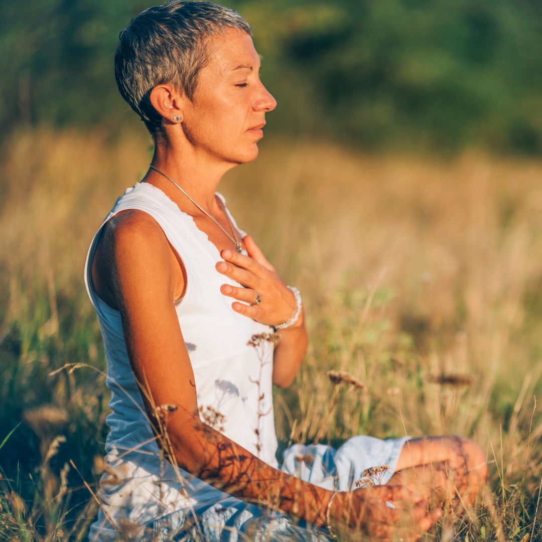 A middle aged woman sits in the grass and meditates with eyes closed and hand on chest.